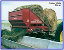 Pulled by tractor along a feed trough, the implement has two big round hay bales being winched into the grinding chamber and the ground up hay being deposited via conveyor into the trough.