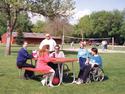 An adult male and female in wheelchairs accessing on one side of the table, its metal frames affixed at each end of both the tabletop and the bench-seat on the other side.