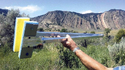 Man's left arm holding up what looks like a giant tong made with handles made of 1-inch square aluminum with a pair of 9-inch long sponges mounted to metal plates on the end. A field & mountains with blue sky in background.