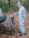 Man in hunting clothes cranking a free-standing hoist with a dead deer on it at the back of a pickup truck.