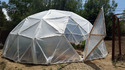 A plastic-covered geodesic-dome sitting in a yard with the dome door open and green trees behind it.